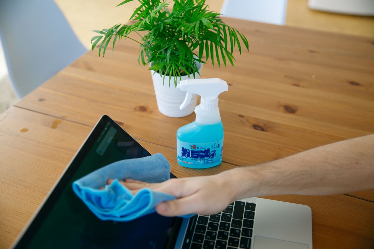 home-img Close-up view of a hand wiping a laptop screen with a blue cloth and spray bottle on a wooden desk.